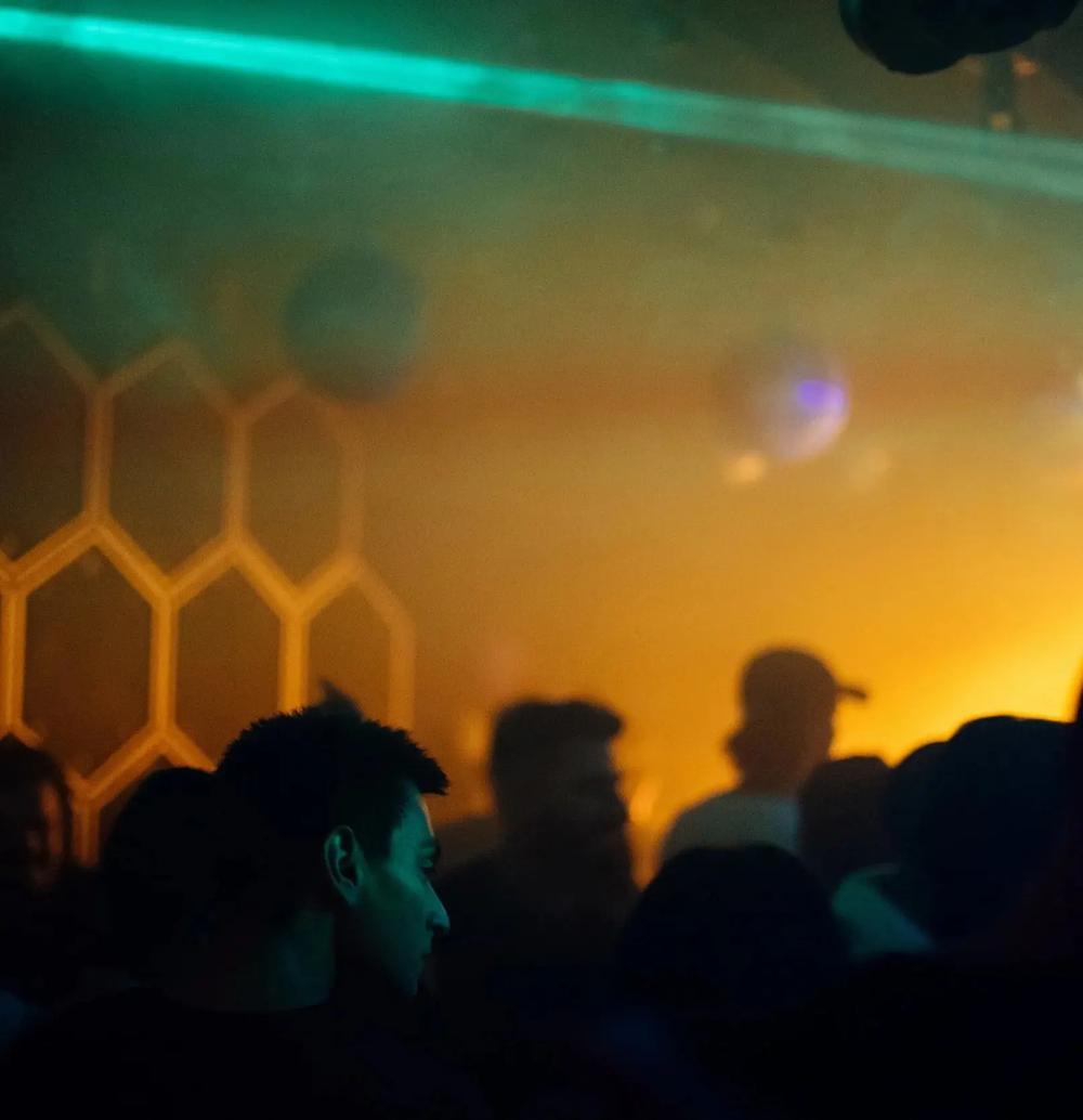 The crowd in silhouette against the hexagon wall, with a teal laser cutting through amber haze